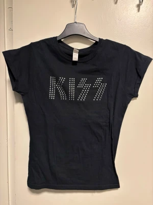 Svart KISS t-shirt med nitar - Cool svart t-shirt med KISS-logga framtill, dekorerad med silvriga nitar som ger en edgy vibe. Modellen är kortärmad och har en klassisk rund hals. Materialet är mjuk bomull som känns skönt mot huden. Perfekt för dig som gillar rock och vill sticka ut.