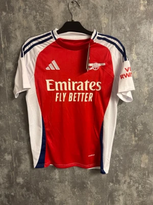 Arsenal fotbollströja Saka - Small - Arsenal matchtröja från Adidas med Saka 7 på ryggen. Tröjan är röd framtill med vita och blå detaljer på ärmarna, samt vita sidor. Tillverkad i lätt och ventilerande polyester, perfekt för fotboll. Emirates och Visit Rwanda-loggor tryckta på bröst och ärm.