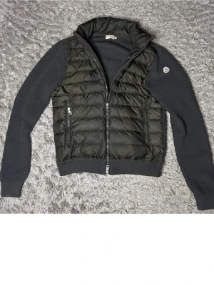 Svart Moncler dunjacka med stickade ärmar - Säljer en svart Moncler dunjacka med quiltad front och ribbstickade ärmar. Jackan har dragkedja framtill, två sidofickor med dragkedja och en hög krage. Klassisk Moncler-logga på ärmen och insida med serietidningsmärke. Perfekt för kyliga dagar.