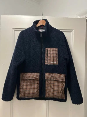 Mörkblå fleecejacka från MC2 St. Barth - Säljer en mörkblå fleecejacka från MC2 St. Barth Après-Ski Collection. Jackan har tre bruna rutiga fickor framtill, hög krage och dragkedja. Materialet är mjuk fleece och detaljerna ger en unik look. Perfekt för kalla dagar.