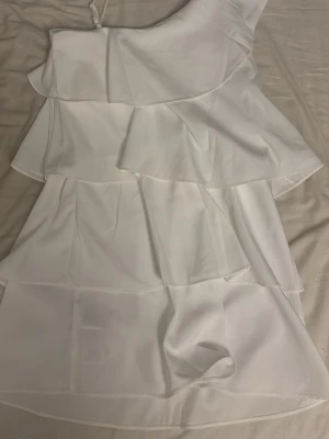 Vit omlottklänning med volanger - Säljer en superfin vit kortklänning med volangdetaljer och omlott-design. Klänningen har en axelbandslös one shoulder-stil och är tillverkad i ett lätt, flowy material som ger en luftig känsla. Perfekt för sommarens alla tillfällen!