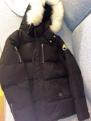 Moose knuckles  - Säljer en svart dunjacka från Moose Knuckles med pälskantad luva och flera dragkedjor. Jackan har en oversized look, coola metallknappar och logga på ärmen. Perfekt för kalla dagar när du vill hålla stilen.