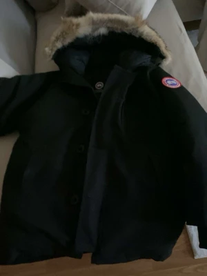 Svart parkas från Canada Goose - Säljer en svart parka från Canada Goose med ikonisk patch på ärmen och stor huva med pälsdetalj. Jackan har knappar framtill och är perfekt för kalla dagar. Klassisk design och riktigt varm, passar dig som vill ha både stil och funktion.