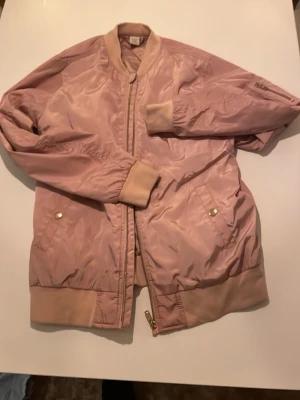 Rosa bomberjacka från Lindex - Säljer en ljusrosa bomberjacka från Lindex med ribbade muddar och krage. Jackan har dragkedja framtill, fickor med tryckknappar och en liten ficka på ärmen. Materialet är glansigt och känns lätt, perfekt för vår eller höst.