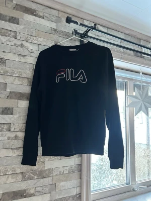 Svart sweatshirt från FILA - Svart sweatshirt från FILA i storlek S. Tröjan har ett stort FILA-tryck i vitt och rött på bröstet och är tillverkad i mjuk bomull. Klassisk passform med långa ärmar och ribbade muddar vid ärmslut och nederkant.