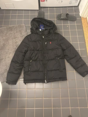 Svart pufferjacka från Polo Ralph Lauren - Svart pufferjacka från Polo Ralph Lauren med huva och dragkedja framtill. Jackan har två sidofickor med dragkedja och den klassiska röda Polo-loggan broderad på bröstet. Perfekt för kalla dagar och har en clean, modern look.