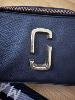 Svart axelväska från Marc Jacobs - Säljer en svart axelväska från Marc Jacobs med guldiga metalldetaljer och logotyp framtill. Väskan har en bred, justerbar axelrem med mönstrad text och dragkedja upptill. Materialet är slitstark syntet med en stilren, boxig form och extra fack på baksidan. Väskan är en 1;1