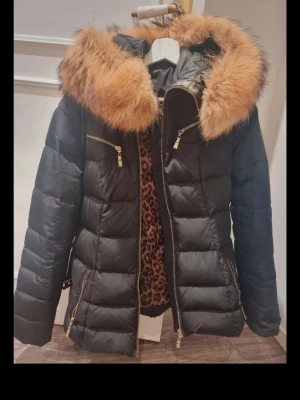 Svart pufferjacka med päls från Hollies - Svart pufferjacka från Hollies med stor fluffig päls på huvan och leopardmönstrat foder. Jackan har guldfärgade dragkedjor på fickor och framtill, samt quiltad design. Perfekt för kalla dagar och riktigt snygg detalj med fodret.