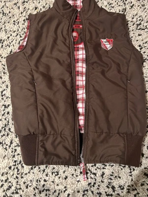 2000s reversible väst  - Säljer en 2000s brun dunväst från Horká med vändbar design. Ena sidan är helbrun med broderat märke och stor rutig siffra 5 på ryggen, andra sidan är rutig i vitt, rött, rosa och brunt. Västen har dragkedja och ribbade muddar nertill. 