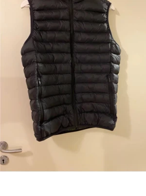 Svart väst  med dragkedja Everest  - Säljer en svart väst  med horisontella quiltade partier och hel dragkedja framtill. Västen har två sidofickor med dragkedja och en klassisk, sportig look. Perfekt att slänga över en hoodie eller tröja när det är kyligt ute. Everest stadium 