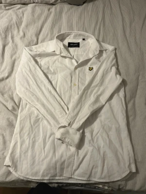 Vit skjorta från Lyle & Scott - Vit skjorta från Lyle & Scott med klassisk krage och gul broderad fågel på bröstet. Skjortan har långa ärmar, knappar framtill och på manschetterna. Tillverkad i mjuk bomull, perfekt för en clean och stilren look.