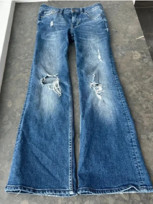 Blå bootcut jeans med slitningar - Första bilderna är från tidigare ägare! Säljer ett par blå bootcut jeans med coola slitningar och hål på knäna och bakfickan. Jeansen har klassisk femficksdesign och är tillverkade i jeansmaterial. Passformen är loose och de har en snygg tvättad look med ljusare partier.