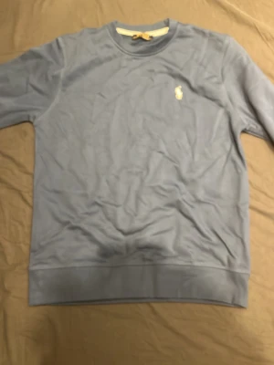 Blå långärmad tröja från Polo Ralph Lauren - Snygg blå långärmad tröja från Polo Ralph Lauren med klassisk rund hals och den ikoniska broderade loggan i vitt på bröstet. Tröjan är tillverkad i mjuk bomull och har ribbade muddar vid ärmslut och nederkant. Perfekt för en chill och stilren look.