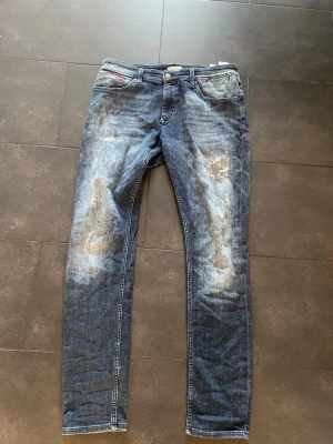 Blåa jeans med slitningar - Säljer ett par jeans i blå tvätt med slitna detaljer och skinny passform. Jeansen har klassisk femficksdesign, dragkedja och knapp i midjan. Materialet är stretchigt jeans och de har coola slitningar på benen för en edgy look.
