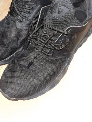 Svarta sneakers från Emporio Armani EA7 - Säljer ett par svarta EA7 sneakers med coolt mönstrad sula och tydlig logga på både plös och häl. Skorna har snörning och är gjorda i syntetmaterial och textil, med en rund tå och platt sula. Perfekta för dig som gillar stilrena och sportiga skor.