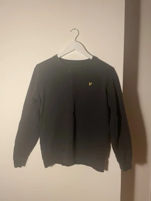 Svart sweatshirt från Lyle & Scott - Svart sweatshirt från Lyle & Scott med klassisk rund hals och broderad gul fågel på bröstet. Använda ganska mycket och inga fläckar. 