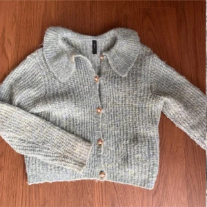 Turkos stickad kofta från YAS - Mysig ljusgrå stickad kofta från YAS med bred krage och snygga guldfärgade knappar framtill. Koftan har långärmad design och ett fluffigt, ribbat material som ger en chill och trendig vibe. Perfekt att slänga på sig när det är lite kyligt ute.