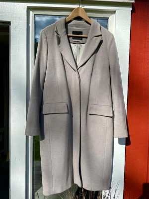 Beige/grå kappa från Oui i ullmix - Snygg beige/grå kappa från Oui med klassisk krage och raka linjer. Tillverkad i en mjuk ullmix med 70% ren ull och 10% Kashmir. Kappan har långa ärmar och två fickor med lock framtill. Perfekt för dig som gillar stilrena och tidlösa plagg. Ena ärmen har en svag fläck, se bild.