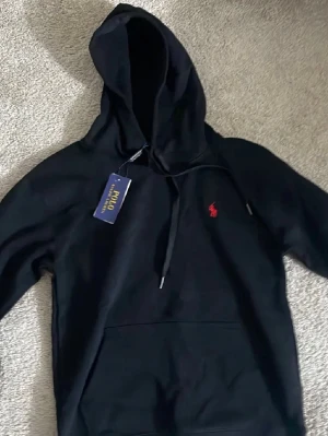 Polo Ralph Lauren hoodie - Svart hoodie från Polo Ralph Lauren med röd broderad logga på bröstet. Klassisk modell med huva, dragsnören och magficka. Tröjan är i fint skick och lappar sitter kvar. Passar bra till vardag och är enkel att matcha.