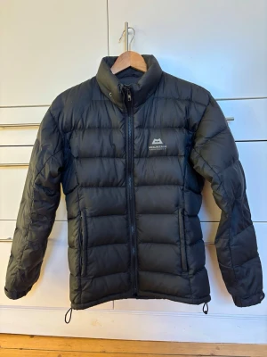 Svart dunjacka i puffermodell från Mountain Equipment, storlek UK 8/XS - Svart pufferjacka i dun från Mountain Equipment. Hög krage. Avtagbar luva (ej på bild men finns!) Logga på bröstet. Justerbar nederkant och ärmslut. Sidofickor med dragkedja. OBS dragkedja trasig i en av fickorna + ett hål på vardera ärm - se bilder. Storlek UK 8/XS