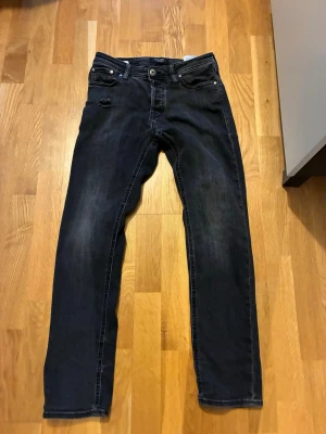 Svarta slim jeans från Jack & Jones - Svarta jeans från Jack & Jones, modell Slim/Glenn. Snyggt slitna med en liten reva vid fickan för extra edge. Klassisk femficksdesign och smal passform som sitter tajt längs benen. Materialet är jeans med en mörk tvätt. Storlek 28/30.