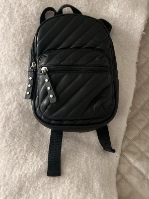 Svart quiltad mini ryggsäck från Zara - Säljer en svart mini ryggsäck från Zara. Har ett litet ytterfack och ett större innerfack. Två justerbara axelband på ryggen och nitar på dragkedjorna.💝 väldigt fint skick 