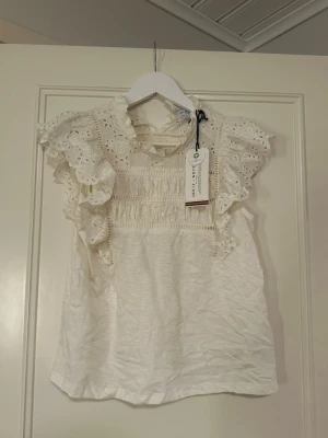 Vit spetsblus från Holly & Whyte - Supersöt vit blus från Holly & Whyte by Lindex med broderade spetsdetaljer och volang vid ärmarna. Blusen knapp i nacken och är ärmlös. Säljer pågrund av att det tyvärr inte kommits tills användning alls. Den är helt ny utan defekter med lappen kvar.