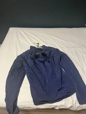 Marinblå Goggle Jacket från C.P. Company - Säljer en marinblå Goggle Jacket från C.P. Company med ikoniska glasögon i huvan. Jackan är tillverkad i lätt och slitstark nylon, har dragkedja framtill, knappdetaljer på fickan och justerbar huva. Perfekt för dig som gillar streetwear och snygga detaljer.