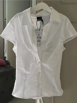 Vit kortärmad skjorta från Gina Tricot - Säljer en fräsch vit skjorta från Gina Tricot i storlek S. Skjortan har korta ärmar, klassisk krage och knappar framtill. Den har snörning baktill för en snygg passform och är gjord i mjuk bomull. Perfekt för dig som gillar stilrena och enkla plagg. 