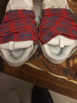 Lanvin sneakers med röda mönstrade band - LÅDA FINNS, köpt secondhand, bra skick. Unika Lanvin sneakers i beige och vitt med tjocka röda och blå mönstrade band istället för vanliga skosnören. Skorna har rund tå, perforerad ovandel och platt sula i gummi. Materialet är en mix av skinn och mocka, vilket ger en lyxig känsla och cool look. Storleken är 43-45 kan vara ganska löst knutna osv 