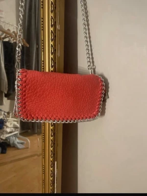 Röd axelväska från Tiamo med kedja - Säljer en snygg röd axelväska från Tiamo med grovt skinnliknande material och silverfärgad kedja som axelrem. Väskan har dekorativt flätad kant och dragkedja upptill. Insidan är svart med en liten innerficka med dragkedja. Perfekt för att lyfta din outfit.