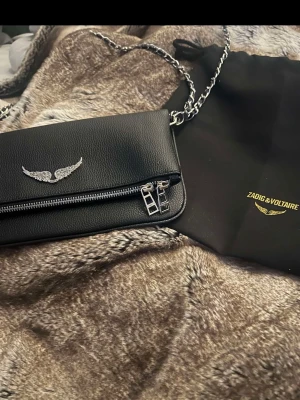 Svart axelväska från Zadig & Voltaire - Säljer en svart axelväska från Zadig & Voltaire i skinn med silverfärgad kedja och detaljer. Väskan har ett snyggt emblem med vingar framtill och dragkedja både fram och bak. Perfekt storlek för det viktigaste och riktigt stilren design. Skriv för fler bilder