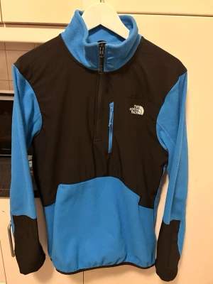 Blå och svart fleece från The North Face - Säljer en blå och svart fleece-tröja från The North Face med half zip och hög krage. Tröjan har en bröstficka med dragkedja och logga på både fram- och baksidan. Perfekt för kyliga dagar och outdoor-aktiviteter. Mycket bra skick, realistiska bud godtas. 
