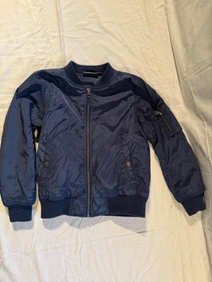 Mörkblå bomberjacka från Lindex - Snygg mörkblå bomberjacka från Lindex med ribbade muddar och krage. Jackan har dragkedja framtill, två fickor med tryckknappar och en extra ficka med dragkedja på ärmen. Materialet är glansigt och känns lätt och bekvämt.