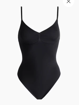 Svart ärmlös bodysuit med smala band - Säljer en svart bodysuit med smala axelband och v-ringad hals. Modellen är tight och har en lätt rynkning vid bysten för extra detalj. Perfekt att styla med jeans eller kjol för en clean look. 