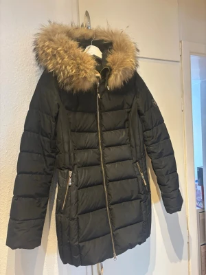 Svart pufferjacka med päls från Hollies - Säljer en svart pufferjacka från Hollies med fluffig beige päls runt huvan. Jackan har guldfärgade dragkedjor, två fickor med dragkedja och snygga quiltade paneler. Perfekt för kalla dagar. Storlek 38