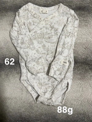 Vit omlottbody med djurmotiv från Newbie - Säljer en söt vit omlottbody från Newbie med ett detaljerat mönster av skogsdjur och växter. Bodyn har långa ärmar och praktiska tryckknappar i grenen. Perfekt för dig som gillar naturinspirerade prints och mjuka material.🌸 Fint använt skick. Övervikbara muddar. Stl 62