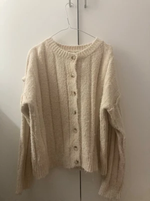 Beige stickad kofta från Daisy Street - Mysig beige kofta från Daisy Street i storlek M. Koftan är stickad med ribbade muddar och har knappar framtill. Den har lång ärm och en loose passform, perfekt för lager-på-lager under kyliga dagar. Materialet är mjukt och fluffigt, vilket gör den extra skön att bära.