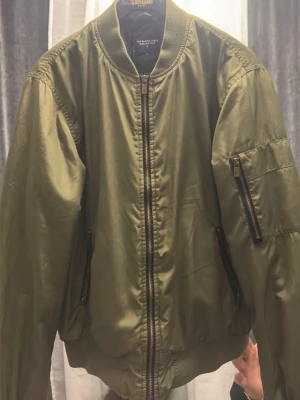 Olivgrön bomberjacka från The Black Label - Snygg olivgrön bomberjacka från The Black Label med klassisk passform. Jackan har dragkedja framtill, fickor på sidorna och en extra ficka med dragkedja på ärmen. Tillverkad i polyester och perfekt för dig som gillar streetstyle.