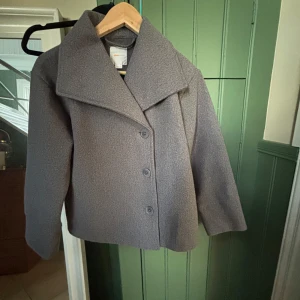 Grå kappa från Gina Tricot - Stilren grå kappa från Gina Tricot med bred krage och tre knappar framtill. Jackan har rak passform och är helfärgad utan mönster, perfekt för dig som gillar en clean look. Materialet känns mjukt och har en ullig struktur.