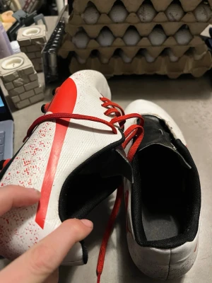 Puma vita och röda fotbollsskor - Säljer ett par fotbollsskor från Puma i vitt med röda detaljer och röd snörning. Skorna har svarta och röda dobbar undertill och ett svart foder. Ovandelen har ett stänkmönster i rött och en stor Puma-logga på sidan. Perfekt för dig som vill sticka ut på planen. En sko saknar snören!