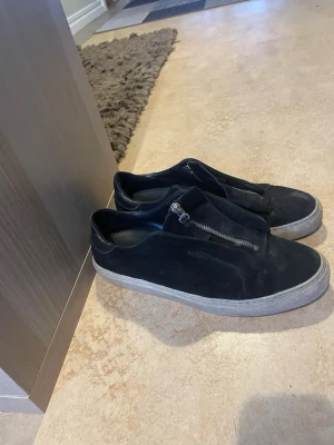 Svarta sneakers från Axel Arigato - Svarta sneakers från Axel Arigato med snygg dragkedja framtill istället för klassisk snörning. Skorna har en rund tå, platt vit sula och är gjorda i mocka. Perfekt för dig som gillar stilrena och moderna detaljer.