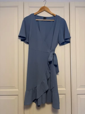 Blå omlottklänning från Vero Moda - Blå omlottklänning från Vero Moda med volangkant och knyt i midjan. Klänningen har korta ärmar och v-ringad hals. Materialet är lätt och har en strukturerad yta.