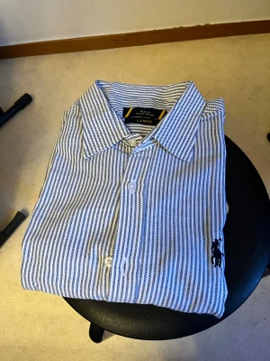 Randig skjorta från Ralph Lauren - Snygg skjorta från Ralph Lauren i storlek Large. Skjortan är vit med smala blå ränder och har klassisk krage, knappar framtill och den ikoniska broderade loggan på bröstet. Perfekt för dig som gillar stilrena och tidlösa plagg.