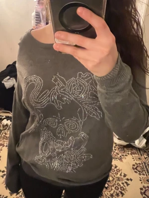 Grå sweatshirt med döskalle och strass - Mysig grå sweatshirt med ett stort döskallemotiv framtill i glittrande strass. Tröjan har lång ärm, avslappnad passform och bred ringning som kan bäras offshoulder för en chill vibe. Perfekt för dig som gillar edgy detaljer och vill sticka ut lite extra.