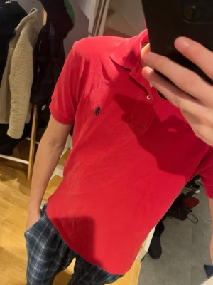 Röd pikétröja från Ralph Lauren - Klassisk röd pikétröja från Ralph Lauren med blå broderad logga på bröstet. Tröjan har krage, korta ärmar och knäppning med två knappar framtill. Perfekt till både jeans och shorts för en avslappnad stil.