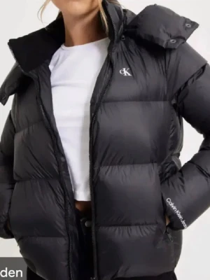 Svart pufferjacka från Calvin Klein - Säljer en svart pufferjacka från Calvin Klein Jeans med huva och dragkedja. Jackan har diskret CK-logga på bröstet och är tillverkad av återvunnen nylon och polyester med dunfyllning. Vattenavvisande material och praktiska fickor med dragkedja.