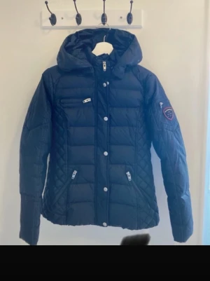 Svart pufferjacka Rockandblue 38 - Svart pufferjacka från Rockandblue med huva, quiltade detaljer och flera dragkedjor. Jackan har ett snyggt märke på ärmen och silverfärgade knappar framtill. Perfekt för kalla dagar och har en modern, normal passform.