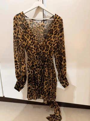 Leopardmönstrad kortklänning Chiquelle - Snygg kort klänning från Chiquelle i leopardmönster med v-ringning och långa ärmar. Klänningen har omlott-design och ett knytband i midjan. Materialet är lätt och luftigt, perfekt för dig som vill sticka ut med en djärv look. Använd 1 gång!