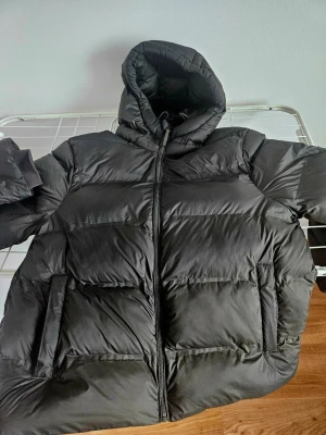 Olivgrön pufferjacka med huva - Säljer en svart pufferjacka med stor, mysig huva och dragkedja framtill. Jackan har breda, quiltade sektioner och två rejäla fickor på sidorna. Perfekt för kalla dagar och riktigt skön att bära. Materialet är syntet och jackan har en loose passform.